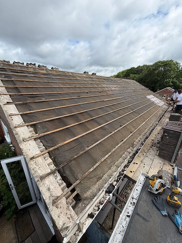 Roof stripped of tiles showing wooden battens with workers and tools on scaffolding at roof edge.