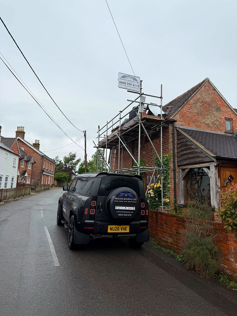 Black SUV parked on a wet street beside a brick house with roofing scaffolding and workers on the roof.
