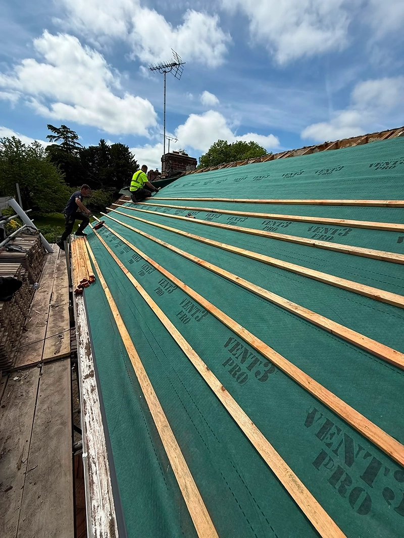 Two workers installing wooden battens on a green roofing underlayment labeled VENT'3 PRO under blue sky.