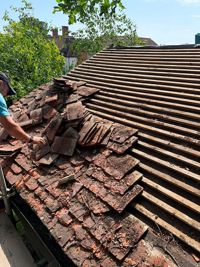 Worker removing old broken roof tiles exposing wooden battens beneath on a sunny day.