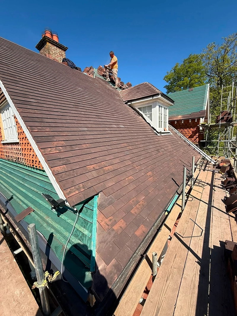 Construction worker laying tiles on a brown rooftop under clear blue sky with scaffolding around.