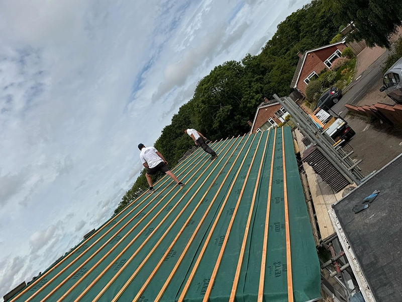 Two workers installing green roofing underlayment with wooden battens on a sloped roof under cloudy sky.
