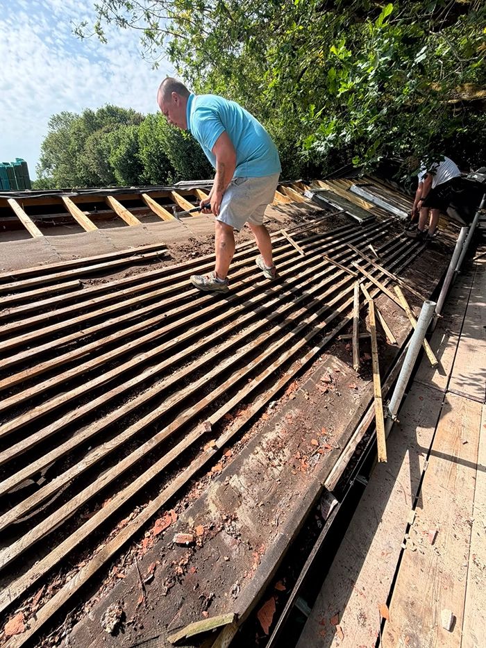 Two men working on a damaged wooden roof under a partly cloudy sky surrounded by trees.