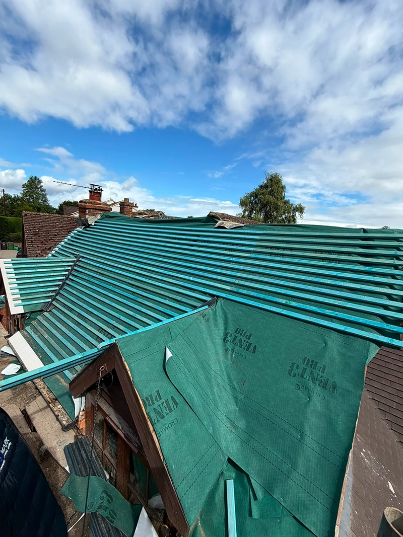 House roof under construction with blue wooden slats and green waterproof membrane under a partly cloudy sky.