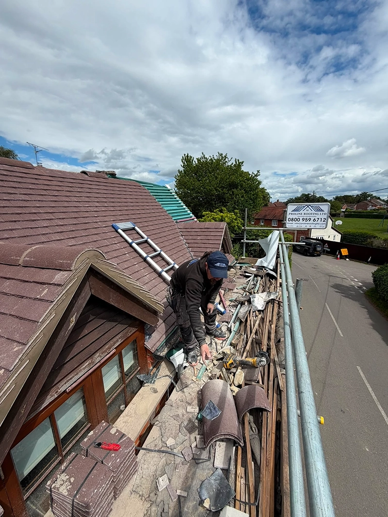 Roofer working on a house roof surrounded by tiles, tools, and debris near scaffolding and a road.