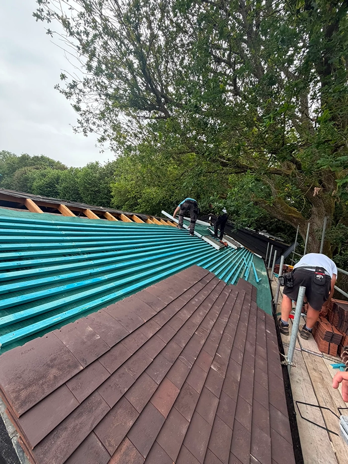 Workers installing brown roof tiles on a sloped teal wooden roof frame with trees in the background.