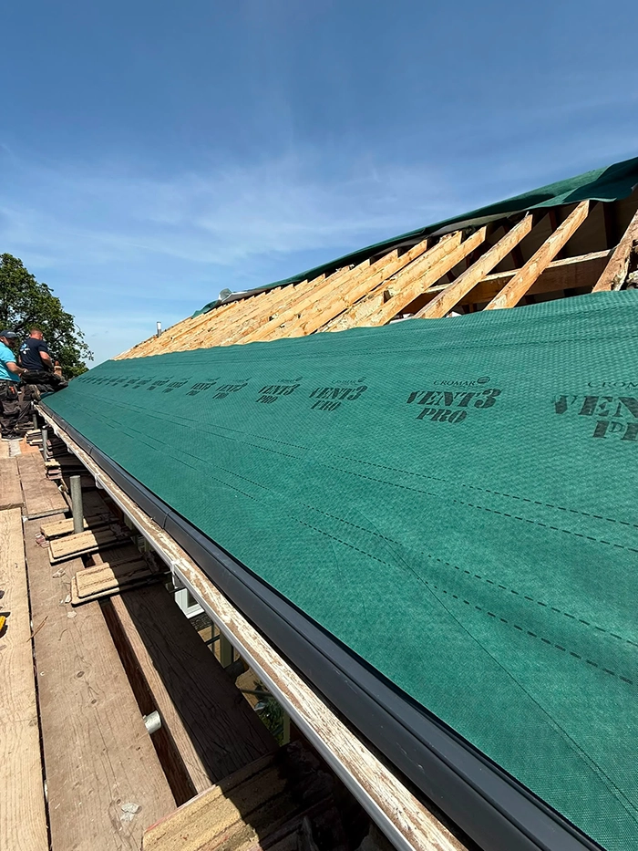 Workers installing green roofing underlayment on wooden roof frame under a clear blue sky.